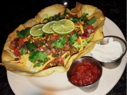 A large taco salad in a crispy shell, topped with ground meat, cheese, lettuce, tomatoes, cilantro, and lime slices, with salsa and sour cream.