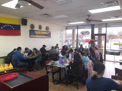 A busy restaurant interior with patrons dining, Venezuelan flag on the wall, and condiments on tables.