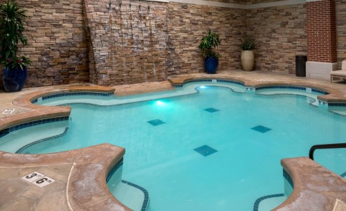 A serene indoor pool with a stone wall, waterfall feature, and blue lighting, surrounded by potted plants.