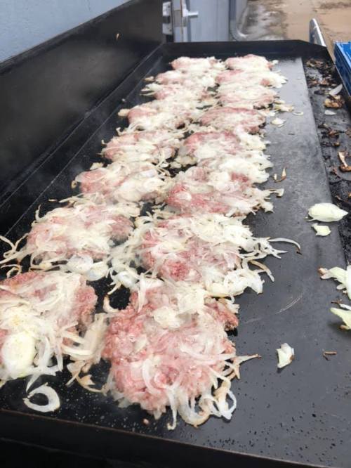 A griddle with multiple patties of ground meat and onions sizzling, releasing steam in an outdoor setting.