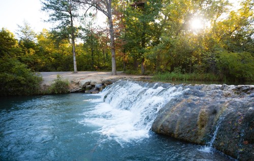 A serene waterfall cascades into a clear blue pool, surrounded by lush green trees and sunlight filtering through.