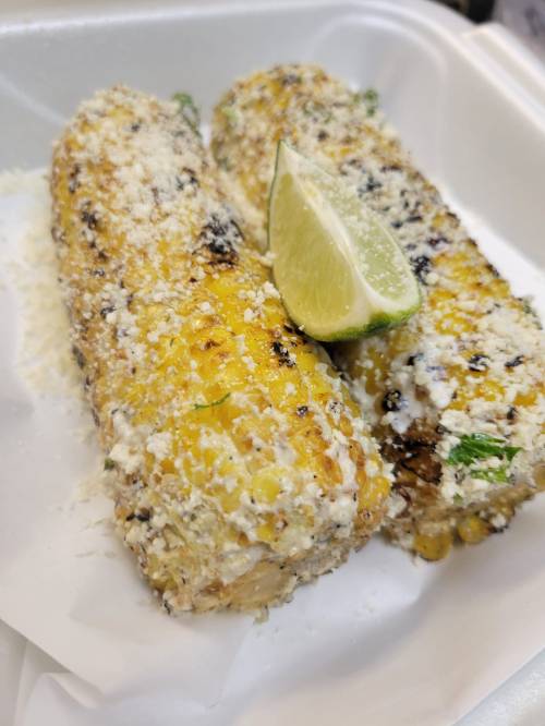 Two grilled corn on the cob topped with cheese and herbs, served with a lime wedge on a white tray.