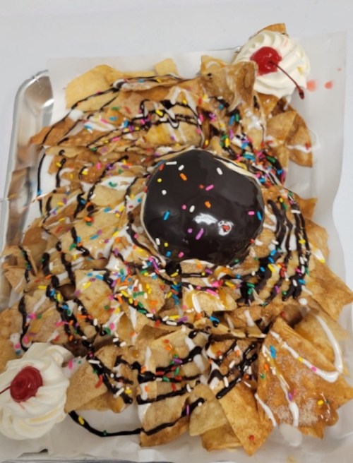 A tray of dessert nachos topped with a chocolate donut, whipped cream, chocolate drizzle, and colorful sprinkles.