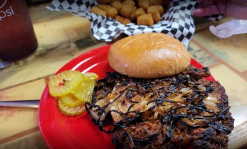A plate with a sandwich topped with grilled onions, pickles, and a side of tater tots in a checkered basket.