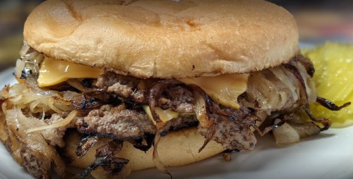 A close-up of a juicy hamburger with melted cheese, grilled onions, and a soft bun, served with a pickle on the side.