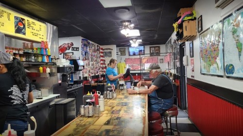 A cozy diner interior with a long counter, patrons seated, and colorful decor on the walls.