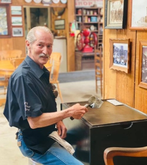 A man with a mustache sits at a table, holding a spatula, in a rustic museum setting with vintage photos on the wall.