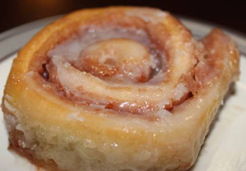 A close-up of a glazed cinnamon roll, showcasing its swirled layers and sticky icing on a white plate.