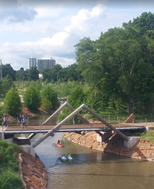 A scenic view of a river with kayakers, a modern bridge, and green trees under a partly cloudy sky.