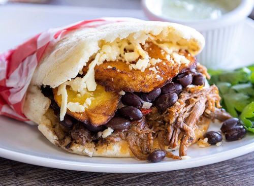 A close-up of a delicious sandwich filled with pulled pork, black beans, fried plantains, and cheese, served with salsa.