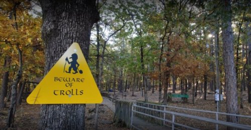 A yellow warning sign reading "Beware of Trolls" is posted on a tree in a wooded area with autumn foliage.