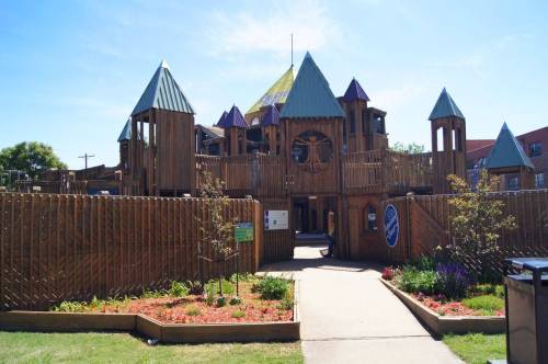 A large wooden playground with towers and a colorful roof, surrounded by gardens and pathways on a sunny day.