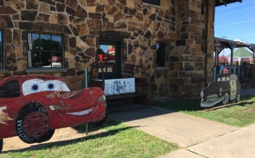 Two wooden cutouts of cartoon cars, one resembling Lightning McQueen, outside a stone building with a grassy area.
