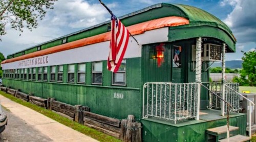 A vintage green train car with an orange roof, featuring an American flag and surrounded by trees and a cloudy sky.