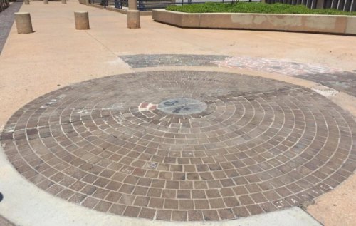 A circular brick pattern on a paved surface, surrounded by concrete and greenery in an outdoor setting.