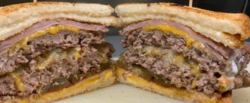 A cross-section of a hearty sandwich with layers of beef patty, ham, cheese, and pickles between toasted bread.