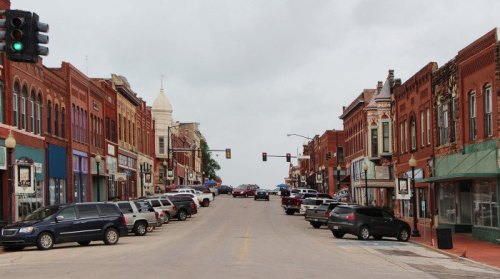 A view of a small town street lined with brick buildings and parked cars, under a cloudy sky. Traffic lights visible.