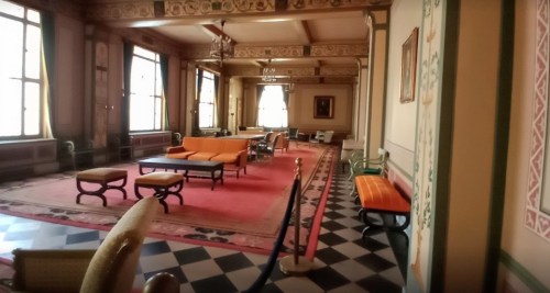 A spacious, elegantly decorated room with orange furniture, large windows, and intricate wall designs.