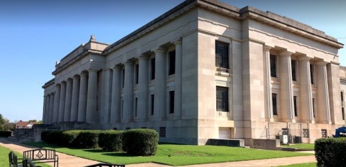 A large, classical-style building with columns, surrounded by green grass and benches in a sunny outdoor setting.