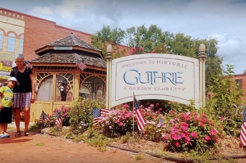 A woman and child walk past a sign welcoming visitors to historic Guthrie, surrounded by flowers and American flags.