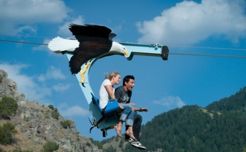 Two people ride a zip line, with a large eagle graphic soaring beside them against a blue sky and mountains.