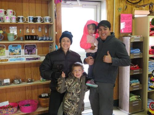 A family of four smiles inside a shop, with two children, one in a pink jacket, and the adults giving thumbs up.