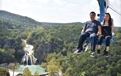 A couple enjoys a scenic chairlift ride with a waterfall and lush greenery in the background.