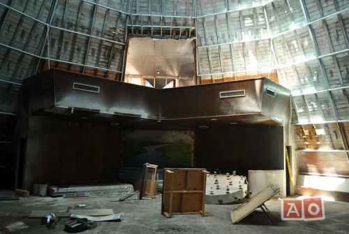 Interior of an abandoned circular building with a high ceiling, featuring scattered furniture and a mural on the wall.