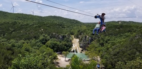 A family enjoys a zip line ride over a lush green landscape with a waterfall and wind turbines in the background.