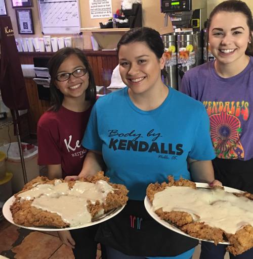 Three smiling waitstaff hold large plates of fried chicken with gravy in a restaurant setting.