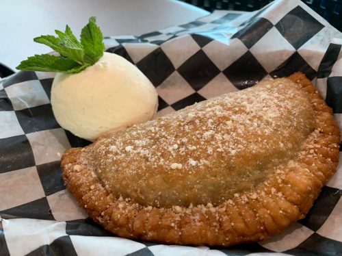 A golden-brown fried pastry with powdered sugar, served with a scoop of vanilla ice cream and a mint leaf on a checkered paper.