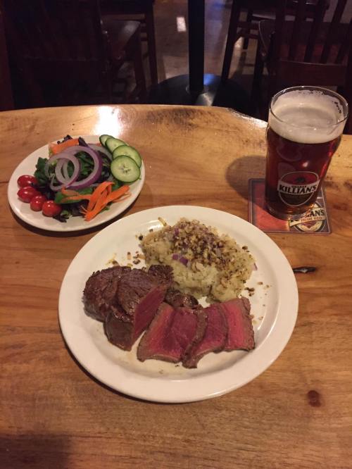 A plate with sliced steak, seasoned rice, a fresh salad, and a pint of beer on a wooden table.