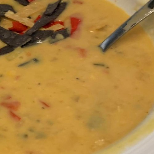 A bowl of creamy soup with red peppers, green herbs, and tortilla strips on top, with a spoon resting beside it.