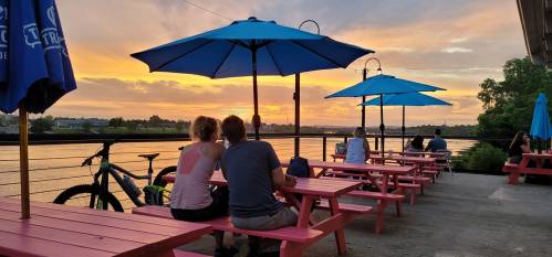 People sitting at pink picnic tables under blue umbrellas, enjoying a sunset by the water. A bike is nearby.