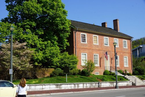 A brick building with a flag, surrounded by greenery, on a sunny day. A person walks by on the sidewalk.