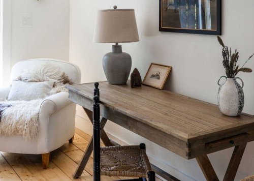 A cozy corner featuring a wooden desk, a lamp, a framed picture, and a plush chair with a throw blanket.