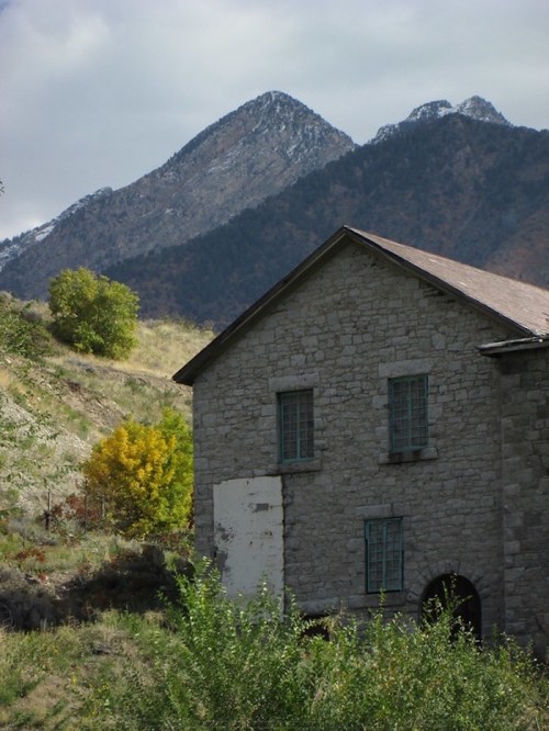 A stone building with green windows, surrounded by trees and mountains in the background under a cloudy sky.