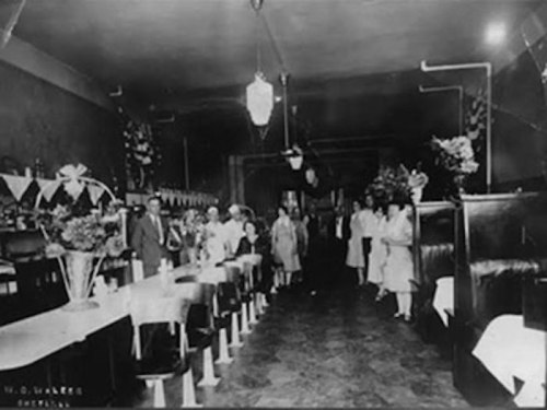 Black and white photo of a vintage restaurant interior with tables, booths, and a group of people standing together.