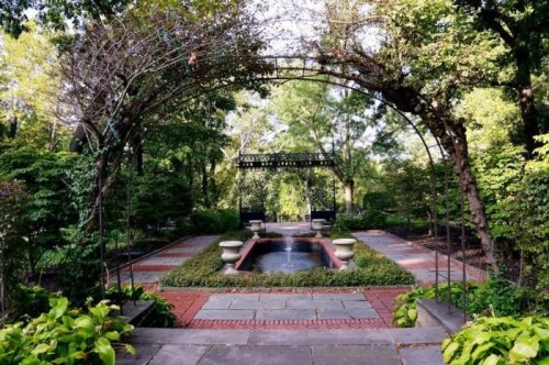 A serene garden with a stone pathway, archway, and a central fountain surrounded by lush greenery.
