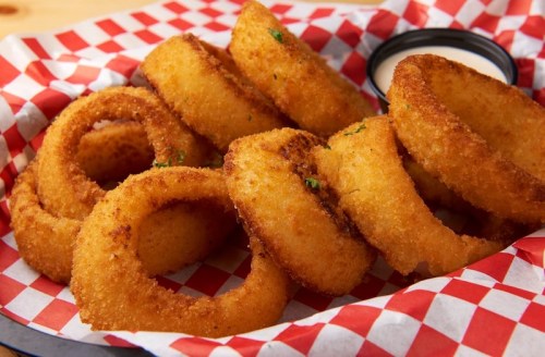 A basket of golden, crispy onion rings served with a side of dipping sauce on a red and white checkered paper.