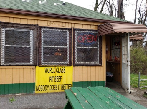 A small, rustic eatery with a green roof, open sign, and a bright yellow sign advertising "World Class Pit Beef."