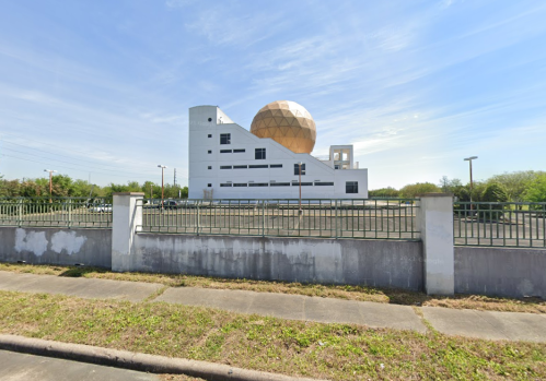 A modern white building with a large golden dome, surrounded by greenery and a fence under a clear blue sky.