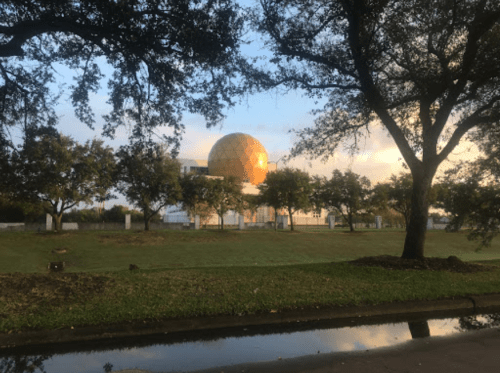 A large golden dome structure surrounded by trees, with a grassy area and a reflective water surface in the foreground.