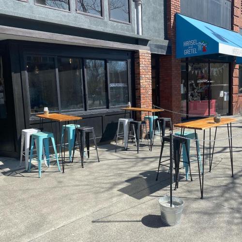 Outdoor seating area with tall tables and stools in front of a restaurant, featuring a blue awning and a bucket on the ground.
