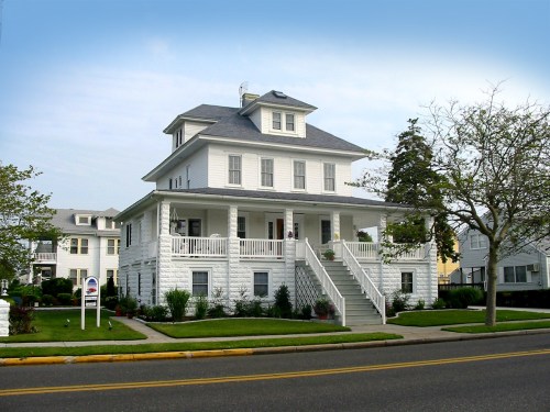 A large white house with a wraparound porch and multiple stories, set on a quiet street with trees nearby.