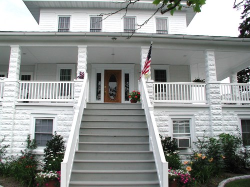 A white two-story house with a large front porch, steps, and colorful flower beds, featuring an American flag.