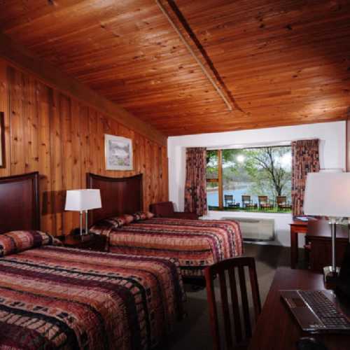 Cozy hotel room with wooden walls, two beds, a desk, and a window overlooking a garden.