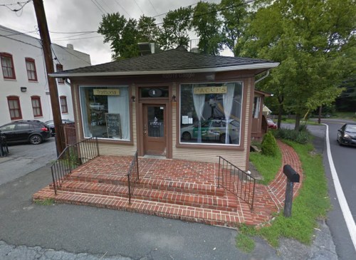 A small restaurant with a brick exterior and a welcoming entrance, surrounded by greenery and parked cars nearby.