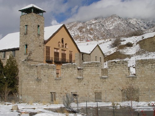 Historic stone building with a tower, surrounded by a wall, set against snow-covered mountains and a cloudy sky.