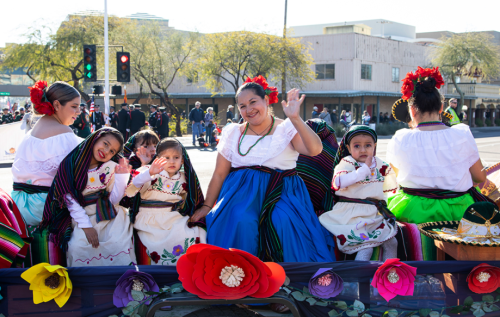 A group of women and children in traditional Mexican attire wave from a colorful float during a parade.